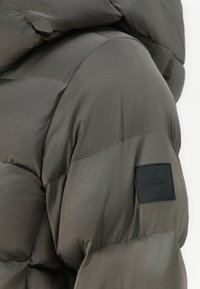 Pufi bunda Calvin Klein v tmavej olivovo zelenej farbe, s lesklou, prešívanou textúrou a čiernou logom nášivkou na rukáve.