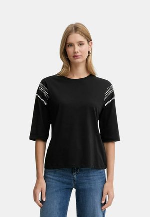 Donna che indossa una maglietta nera a maniche corte con borchie decorative argentate sulle spalle, abbinata a jeans blu, in piedi davanti a uno sfondo uniforme.