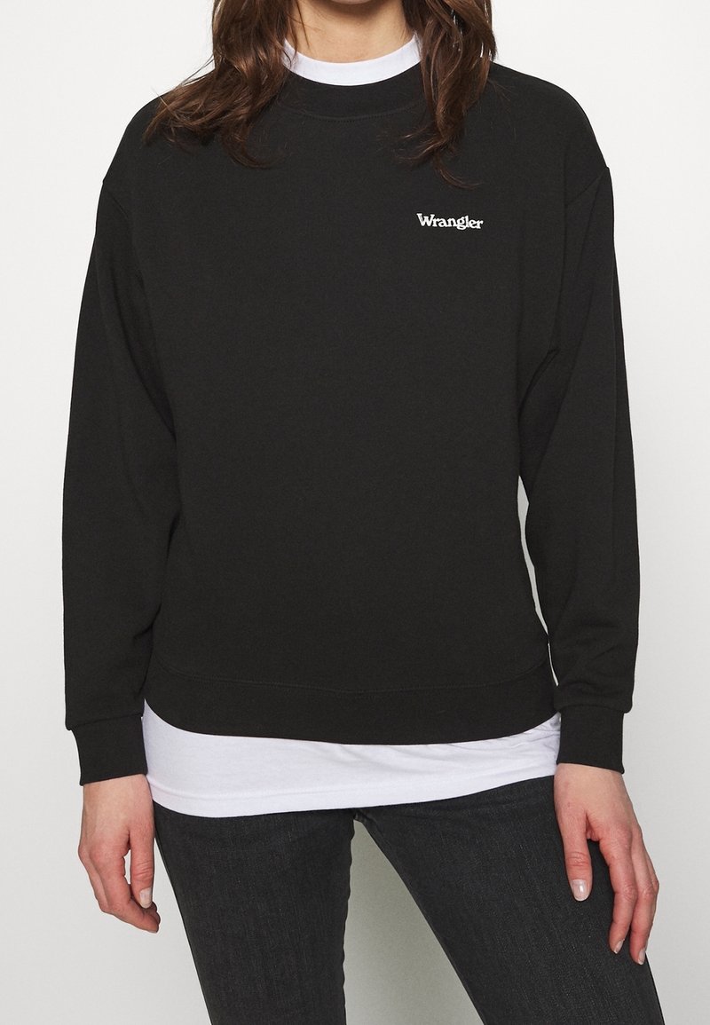 Personne portant un sweat-shirt noir Wrangler par-dessus une chemise blanche et un jean foncé, debout devant un fond blanc, les bras détendus le long du corps.