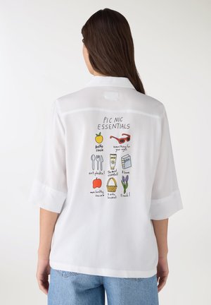 Donna che indossa una camicia bianca con illustrazioni colorate di accessori da picnic e testo sul retro, abbinata a jeans azzurri.