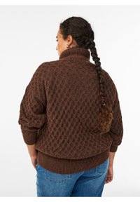 Bruine gebreide trui met een hoge kraag, voorzien van een textuurpatroon en geribde manchetten en zoom. Gecombineerd met blauwe denim jeans.