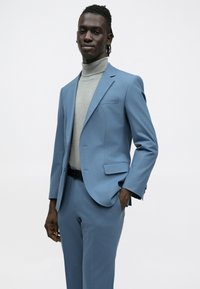 Világoskék öltönyzakó bevágott hajtókkal, szürke garbóval párosítva. Simább anyag, testre szabott szabás és finom zsebrészlet.