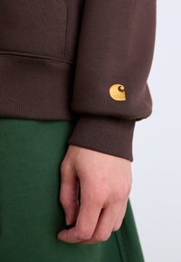 Brown sweatshirt with ribbed cuffs, featuring a distinctive gold logo on the sleeve. A green garment is visible beneath.