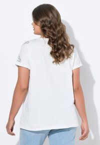 Camiseta blanca de manga corta con puños enrollados, que presenta acentos gráficos negros en los hombros. Textura suave, corte relajado y dobladillo redondeado.