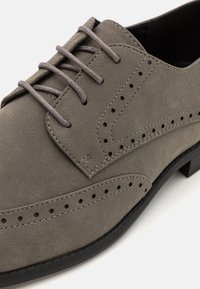 Chaussure habillée en daim gris à lacets avec perforations de style brogue et semelle noire, présentée en gros plan.