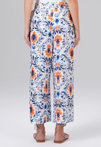 Witte bloemen wijde broek met oranje en blauwe patronen. Elastische tailleband, losse pasvorm en enkellange ontwerp. Platte sandalen zichtbaar.