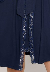 Granatowa sukienka z koronkowym wykończeniem wzdłuż przedniego rozcięcia. Miękki, gładki materiał o płynnej formie oraz delikatne koronkowe detale w falbanki.