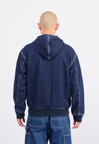 Mørkeblå denim hoodie-jakke med afslappet pasform, synlige sømme, elastiske manchetter og talje. Har en hætte med snore.