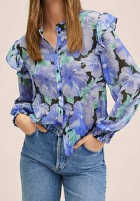 Blouse fleurie en tissu transparent avec des fleurs bleues et vertes sur un fond noir. Conception à boutons sur le devant avec manches à volants et poignets élastiques.