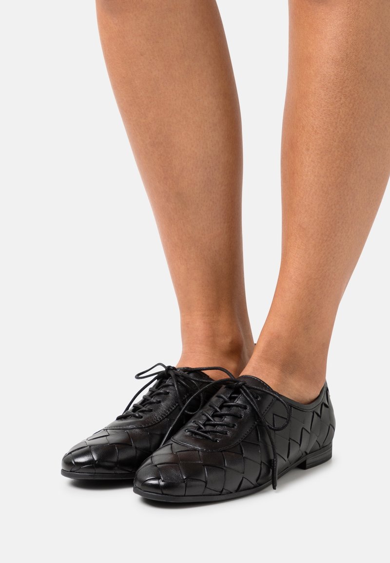 Chaussures à lacets en cuir tissé noir portées sur des pieds avec jambes nues, présentées sur un fond gris clair uni.