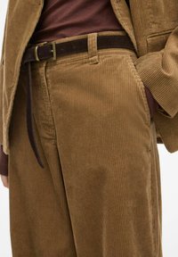 Bruine corduroybroek met verticale ribbels, voorzien van een bruine riem met een rechthoekige metalen gesp. Gecombineerd met een donker maroon top.