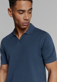 Kortarnik, marineblauwe polo trui van zacht breigoed. Heeft een kraag en een V-vormige opening aan de voorkant.