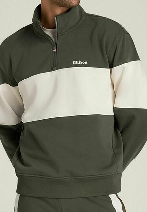 Uomo che indossa un pullover a blocchi di colore verde oliva e crema con mezza cerniera e logo "Wilson" sul petto, mani nelle tasche.