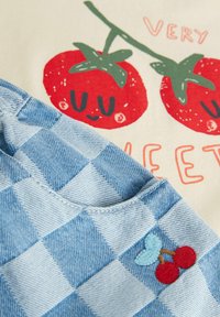 Tissu en denim à carreaux bleu clair avec un design de cerise brodé ; le fond crème présente un graphique de deux tomates rouges souriantes et du texte.