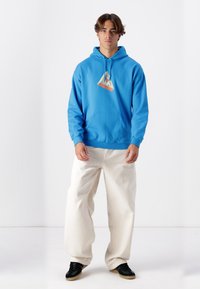 Blue cotton hoodie with a triangular graphic featuring orange and yellow accents. Paired with cream wide-leg pants and black sneakers.