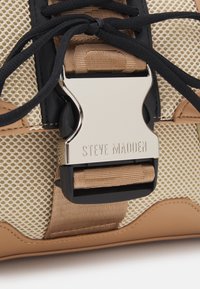 Bolso mensajero en tono marrón y negro con panel de malla beige, que presenta un prominente cierre de hebilla plateada marcado "STEVE MADDEN". Superficie texturizada y diseño en capas.