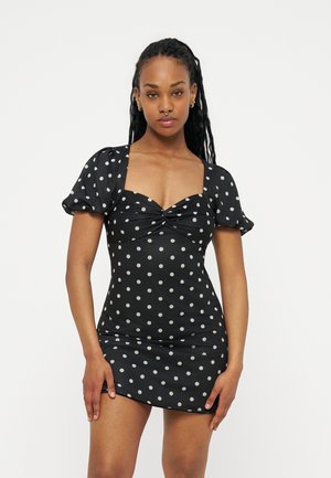 Vrouw met lang, gevlochten haar, gekleed in een zwarte minijurk met witte stippen, pofmouwen en een gedraaide voorkant, staand tegen een witte achtergrond.