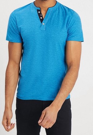 Mężczyzna ubrany w niebieską koszulkę typu henley z krótkim rękawem i czarną plisą na guziki, w połączeniu z czarnymi spodniami, stoi na tle jednolitego białego tła.