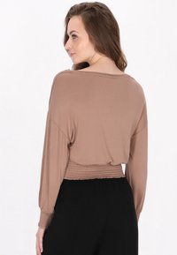 Top corto marrone a maniche lunghe realizzato in materiale morbido con scollo ampio e orlo arricciato. Abbinato a pantaloni neri a vita alta.