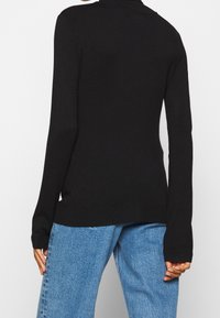 Czarny dzianinowy sweter z długimi rękawami i wysokim dekoltem, o dopasowanym kroju i ściągaczami przy mankietach, zestawiony z jasnoniebieskimi dżinsami.