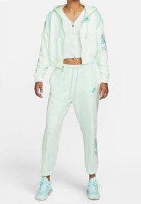 Jasnozielona przycięta bluza z kapturem w zestawie z dopasowanymi dresami. Miękka tkanina, zapinana na zamek błyskawiczny z przodu, ozdobiona haftowanymi detalami. Biały sneakersy dopełniają stylizację.