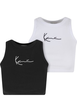 Vrouw die een wit cropped tanktop, losse zwarte broek en witte sneakers draagt, staat met één hand in haar zak en kijkt naar de zijkant.