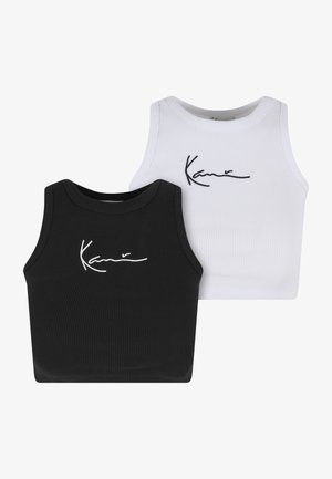 Femme portant un débardeur court blanc, un pantalon noir ample et des baskets blanches, debout avec une main dans la poche, regardant sur le côté.