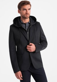 Schwarze Jacke mit Kapuze, mit einem Reißverschluss vorne und einer schmalen Passform. Glatte Textur und minimalistisches Design, ergänzt durch ein graues Hemd darunter.