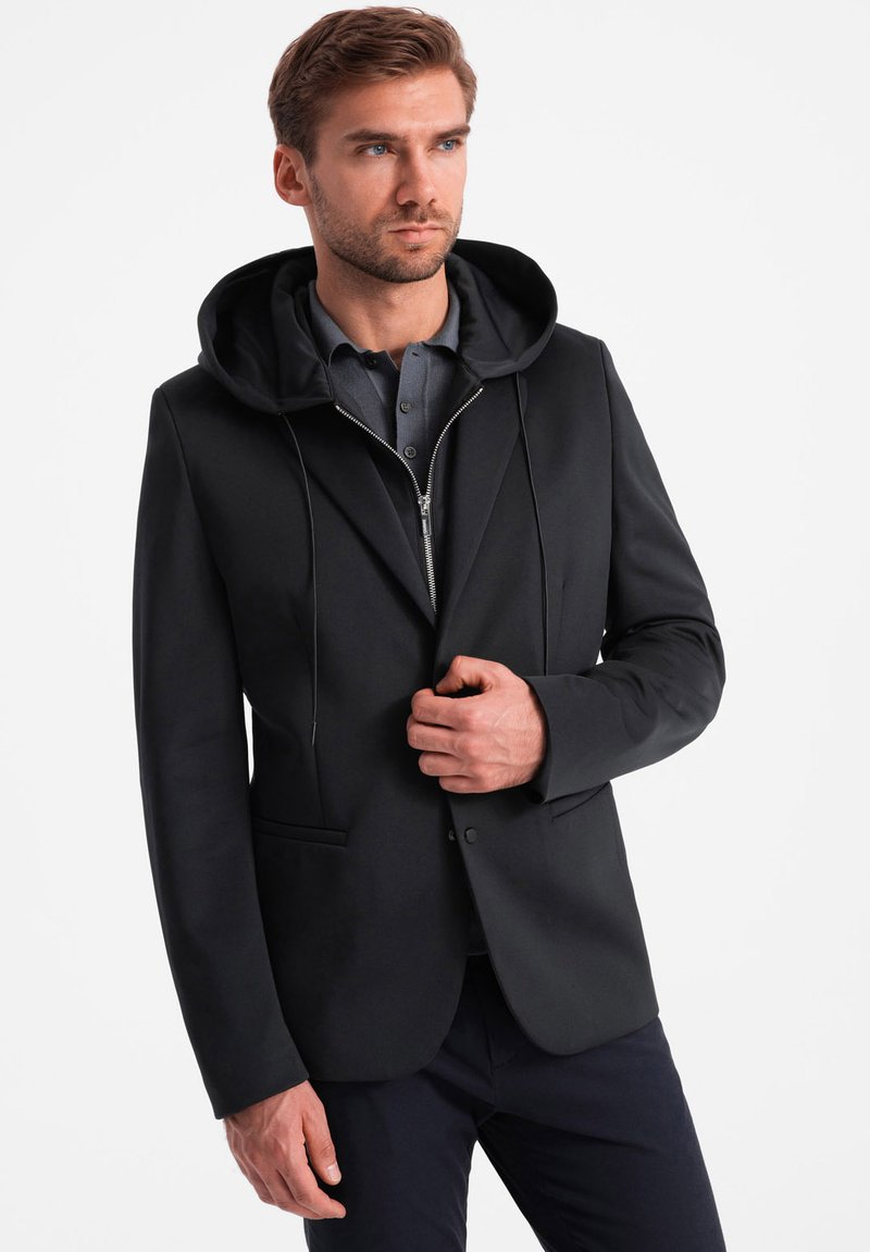 Giacca nera con cappuccio, dotata di una chiusura zip frontale e una vestibilità slim. Texture liscia e design minimalista, completata da una camicia grigia sottostante.