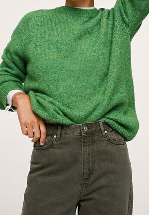 Osoba w zielonym dzianinowym swetrze wsuniętym w wysokowalne, wyblakłe czarne dżinsy, z jedną ręką spoczywającą blisko kieszeni.