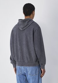 Sudadera de algodón cepillado en color gris, con capucha ajustable y puños acanalados. Presenta un corte relajado y una textura sutil, llevada sobre jeans de color azul claro.