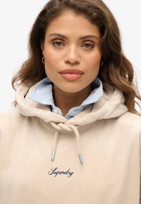 Beżowa bluza z sztruksu z jasnoniebieską koszulą z kołnierzykiem pod spodem, z trokami do regulacji i haftem "Superdry" w ciemnoniebieskim kolorze.