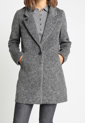Femme portant un manteau mi-long gris en laine avec des revers crantés, deux poches devant et une fermeture à un bouton sur un polo gris.