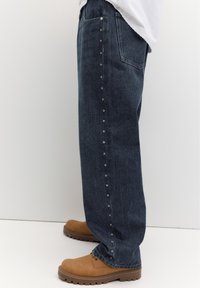 Jean bleu foncé avec de petites décorations en forme d'étoile le long de la couture latérale, porté avec des chaussures marron robustes et un haut blanc.