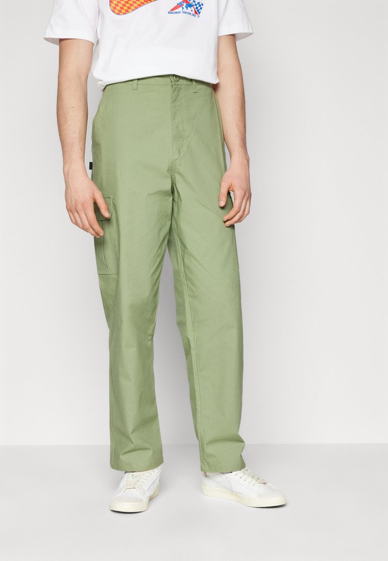 Pantalon cargo vert, coupe décontractée, avec un motif texturé et des poches latérales, associé à des baskets blanches et un t-shirt graphique.