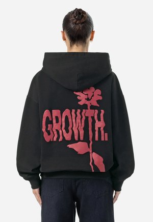 Osoba z włosami związanymi z tyłu, nosząca czarną bluzę z kapturem z dużym różowym kwiatem i napisem „GROWTH.” na plecach.