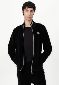 Giacca in corduroy nera con zip bianca e rifinitura color colletto, caratterizzata da un piccolo logo "OPEN ERA" sul lato sinistro del petto. Indossata sopra una camicia nera.