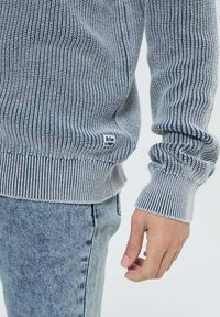 Pull en tricot bleu et gris clair avec un ourlet et des poignets côtelés, arborant un petit logo noir en bas à gauche.
