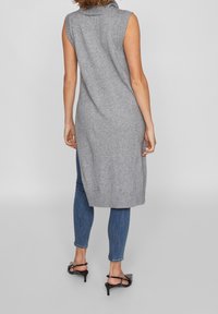 Grå ärmlös stickad tunika med hög krage, sidofickor och ribbad fåll, kombinerad med blå skinny jeans och svarta klacksandaler.