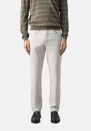 Hombre que lleva pantalones beige claro, mocasines negros y un suéter verde oliva con rayas blancas horizontales, de pie frente a un fondo liso.