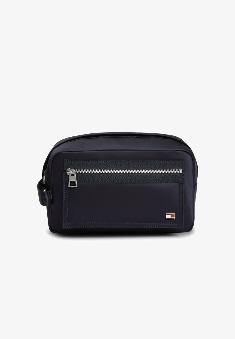 Petite trousse de toilette noire compacte avec poche zippée à l'avant, poignée latérale, et petit logo rectangulaire rouge, blanc et bleu dans le coin inférieur droit.