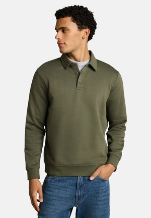 Mann trägt ein olivgrünes langärmeliges Poloshirt über einem grauen Unterhemd und blauen Jeans, schaut nach links mit einer Hand in der Tasche.