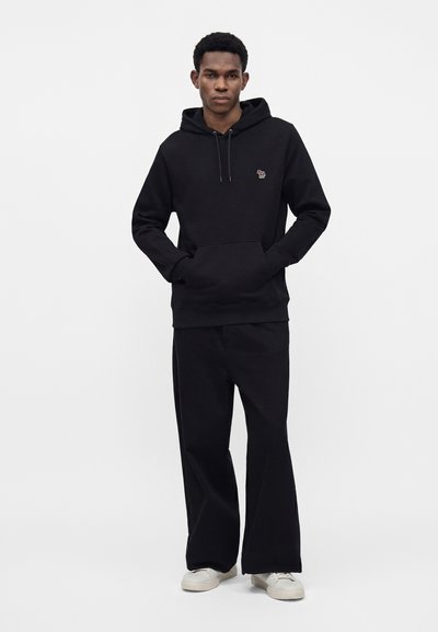 Jeune homme debout, les mains dans les poches d'un sweat à capuche noir, portant un pantalon noir à jambes larges et des baskets blanches sur un fond uni.