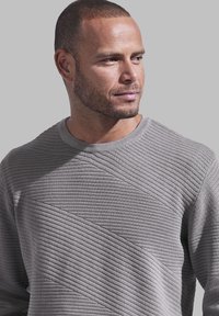 Grauer strukturierter Pullover mit diagonaler Rippstruktur, Rundhalsausschnitt und kontrastierenden glatten Akzenten. Das Material wirkt weich und bequem.