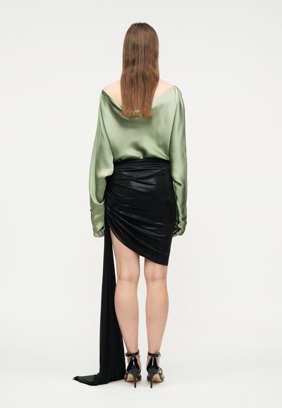 Γυναίκα με μακριά καστανά μαλλιά φοράει πράσινο σατέν μπλουζάκι με ανοικτούς ώμους και μια μαύρη ασύμμετρη φούστα με μακριά, χαλαρά κρεμασμένη ουρά, στέκεται με την πλάτη γυρισμένη.