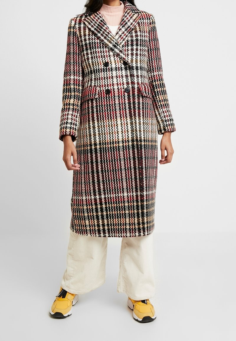 Dvojřadý kabát v barevném kostkovaném vzoru, s klopovým límcem, dlouhými rukávy a střední délkou. Nositý přes béžové kordové kalhoty.