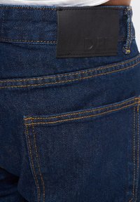 Dark blue denim jeans featuring a black leather patch labeled "DEF" on the back waistband, with visible orange stitching details.