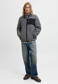 Grijze fleeced hoodie met een zwart paneel, ritssluiting en twee voorkeurszakken, gecombineerd met een geruit overhemd en lichtblauwe verwasemde jeans. Bruine schoenen.