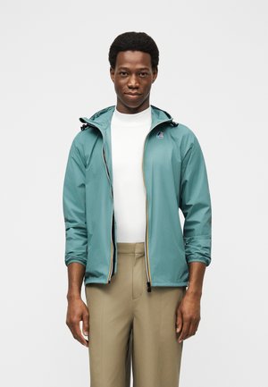 Mann iført teal hettejakke over hvit skjorte og beige bukser, stående mot en enkel lys bakgrunn.