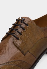 Καφετί δερμάτινο παπούτσι για φόρεμα με μαύρα κορδόνια, διαθέτοντας υφασμάτινα και λείες επιφάνειες σε κοντινή λήψη.